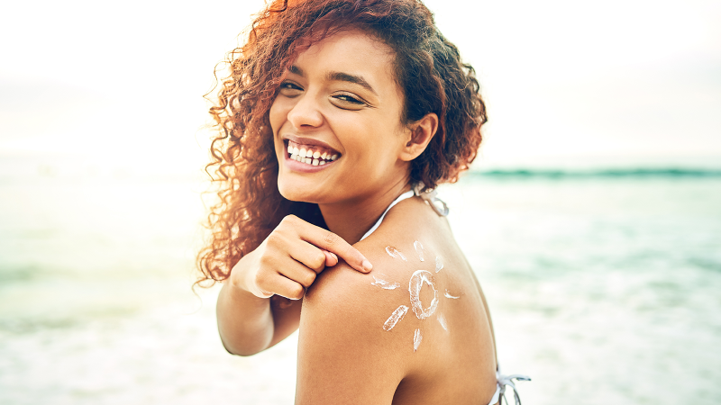 young woman on at the beach in a bikini, looking at the camera and smiling while pointing at a stylized drawing of a sun on her shoulder made with sunscreen 