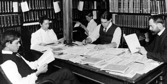 Four men and one woman seated in a library with journals spread on a table in front of them