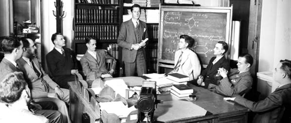 Nine men sitting around a conference table and one man standing in front of a backboard showing chemical diagrams