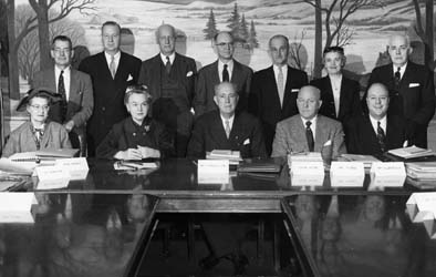 Group picture of three women and nine men gathered behind a conference table with stacks of paper in front of them