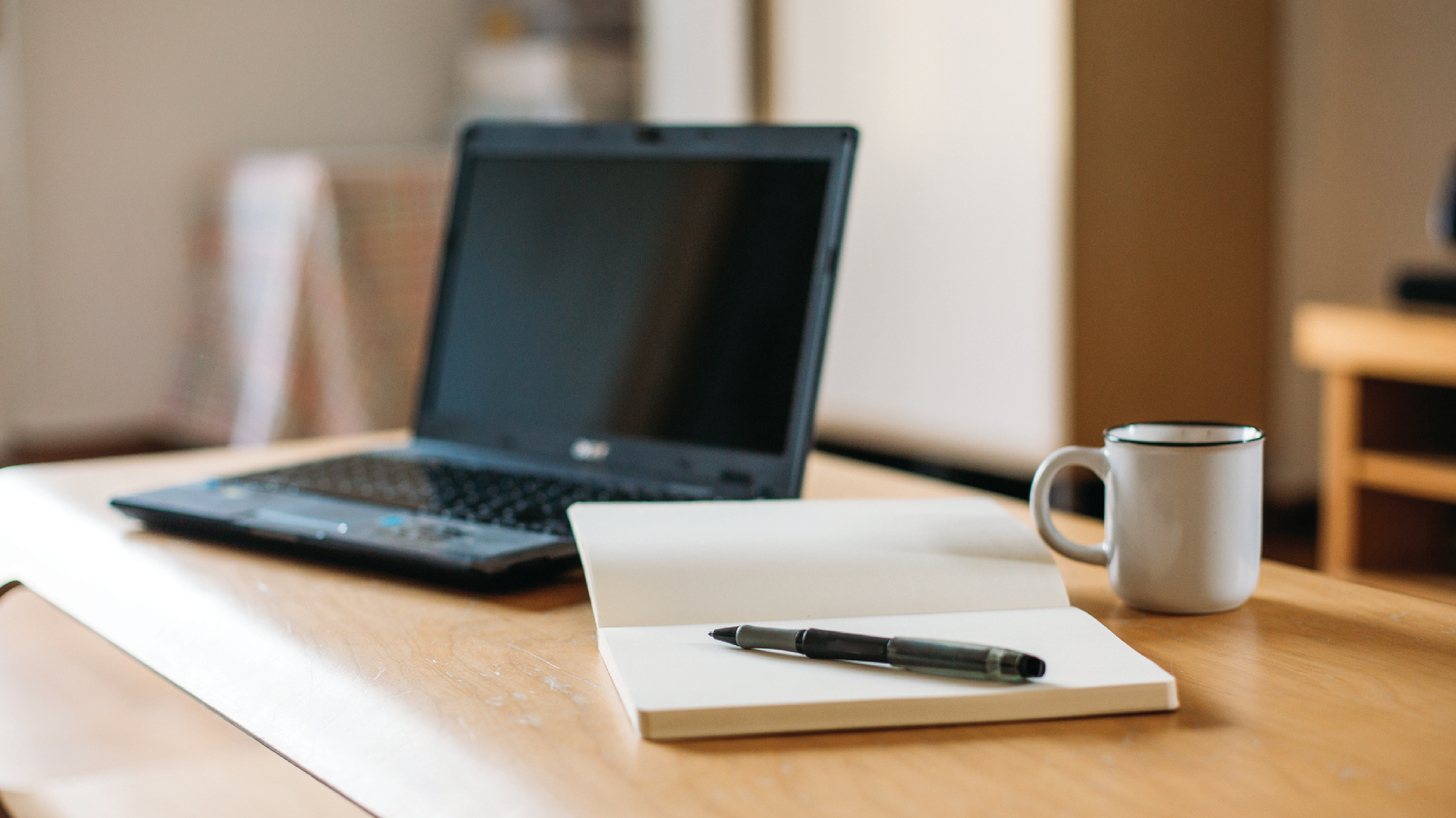 A desk with a laptop computer, notebook, pen, and mug.