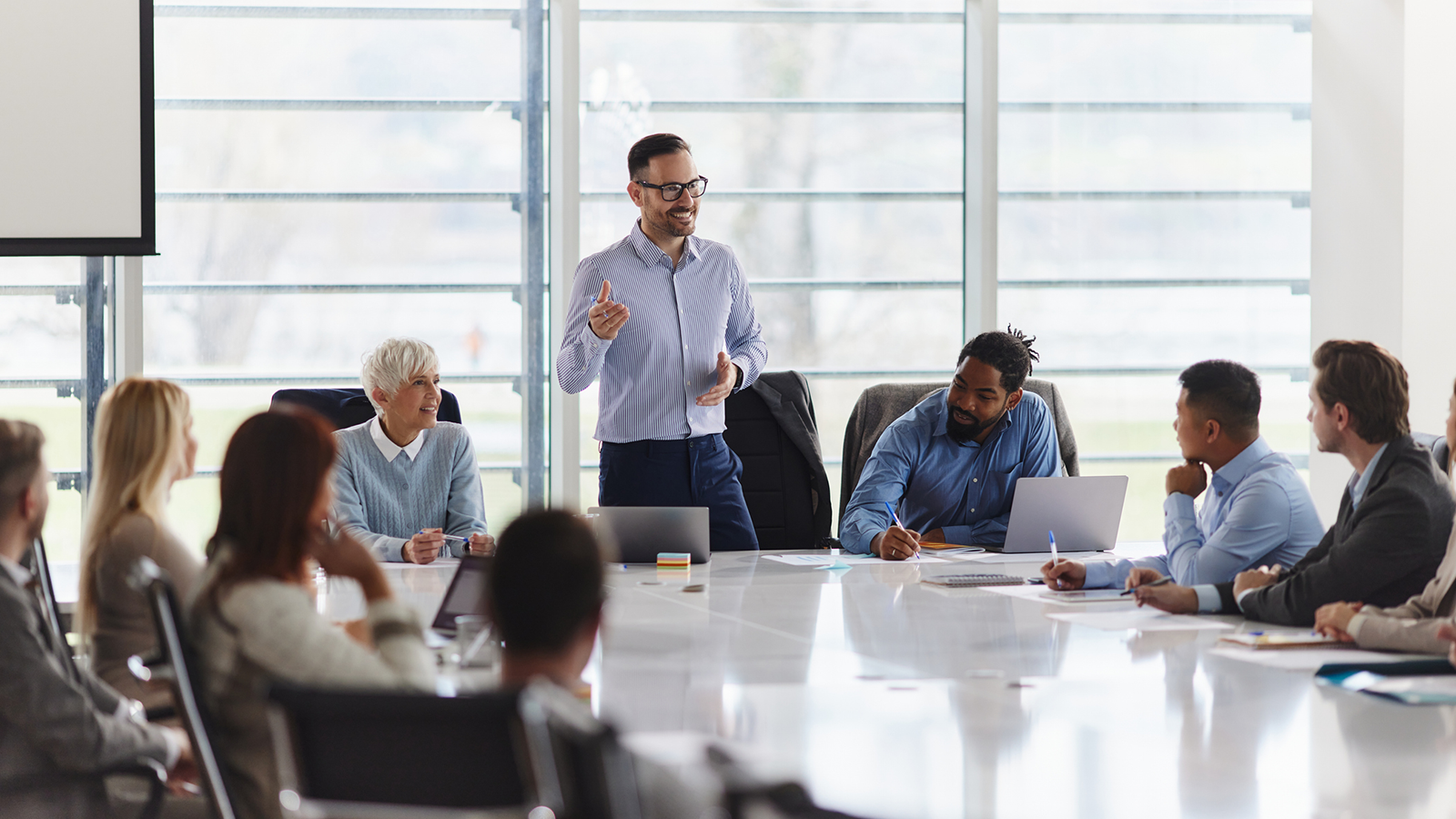 Man standing up and speaking in front of group of people sitting around a table