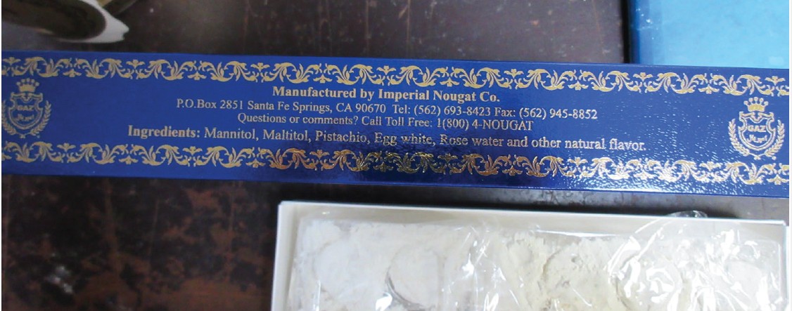 A close-up photograph of the side panel of a blue and gold decorative box. The panel features ornate gold scrollwork borders along the top and bottom edges, with the Imperial Nougat Co. logo on each end. The text reads: "Manufactured by Imperial Nougat Co. P.O. Box 2851 Santa Fe Springs, CA 90670 Tel: (562) 693-8423 Fax: (562) 945-8852. Questions or comments? Call Toll Free: 1(800) 4-NOUGAT. Ingredients: Mannitol, Maltitol, Pistachio, Egg white, Rose water and other natural flavor." A partially visible whit