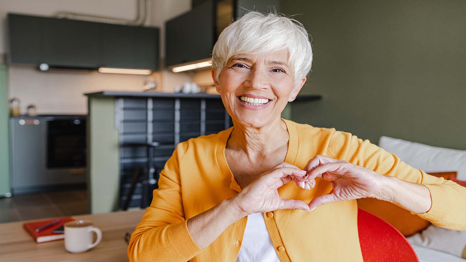 Older Caucausian woman smiling at camera making a heart smbol with her fingers