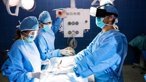 A surgeon wearing blue scrubs and using a virtual reality headset to operate on an individual. He is aided by two nurses wearing blue scrubs. 