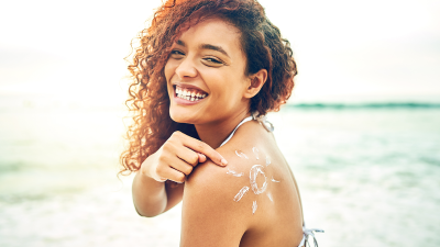 young woman on at the beach in a bikini, looking at the camera and smiling while pointing at a stylized drawing of a sun on her shoulder made with sunscreen 