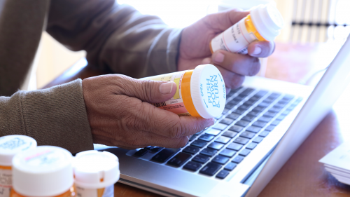 Photo of senior citizen reading labels on prescription drug bottles while at his laptop computer.