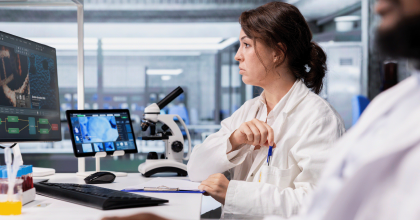 A scientist in a lab coat uses a digital tablet to analyze a colorful 3D cellular model while collaborating with a colleague.