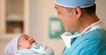 A male doctor in blue surgical scrubs and a cap looks down lovingly at a newborn baby he is holding. The baby, swaddled in a striped blanket and cap, looks up at the doctor.