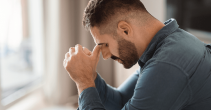 A man in a blue shirt with his eyes closed, leaning forward and pinching the bridge of his nose as if in pain or from stress.