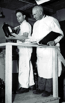 Man in a white lab coat checking an open notebook and the label of a large jar being held by a second man wearing a coverall.