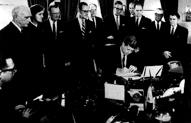President Kennedy seated at a desk, signing a document, surrounded by a group of ten men and one woman
