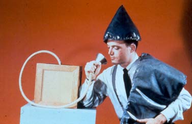 A man is sitting down wearing a cone shaped hat, holding a sash-like device over his left side, and he is holding a cone shaped device to his mouth.