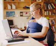 Woman sitting at a desk working on a laptop computer, with a baby wrapped up against her.